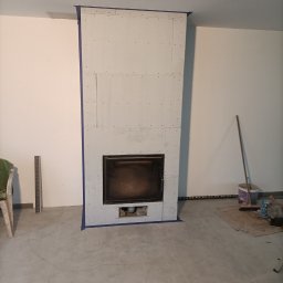 RO-STA Firma Ogólnobudowlana Starosta Roman - Wnętrze z kominkiem w trakcie obudowy płytami kartonowo-gipsowymi, zabezpieczonymi niebieską taśmą malarską, widoczne narzędzia i pozostałości po pracy.
