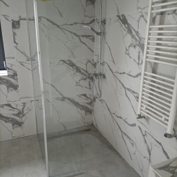 RO-STA Firma Ogólnobudowlana Starosta Roman - Kabina prysznicowa typu walk-in z przezroczystego szkła, zamontowana w łazience wykończonej płytkami imitującymi marmur, z widoczną baterią prysznicową i grzejnikiem drabinkowym.