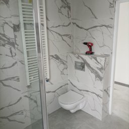 RO-STA Firma Ogólnobudowlana Starosta Roman - Nowoczesna łazienka z marmurowymi płytkami, wiszącą toaletą i szklaną kabiną prysznicową w trakcie wykańczania; na półce widoczna wiertarka akumulatorowa.