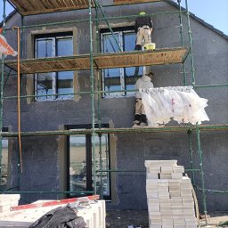 RO-STA Firma Ogólnobudowlana Starosta Roman - Ocieplanie elewacji domu jednorodzinnego: pracownicy na rusztowaniu podczas montażu styropianu, widoczne worki z klejem, narzędzia i materiały budowlane.