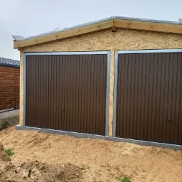 Montaż dwuskrzydłowej, brązowej bramy garażowej w surowej konstrukcji z płyt OSB, widoczne drewniane elementy więźby dachowej zabezpieczone folią budowlaną, teren wokół w stanie przygotowania.