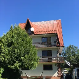 Trzypiętrowy dom z pomarańczowym, metalowym dachem i balkonami, widziany w słoneczny dzień. Na dachu widoczna antena satelitarna. Na pierwszym planie duży, zielony krzew i częściowo widoczny hamak.