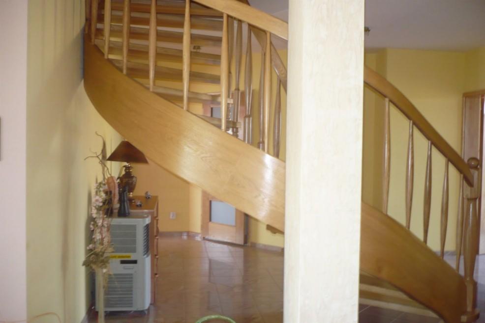 Drewniane schody kręcone z balustradą i dekoracyjną kolumną, widok na wnętrze z terakotą na podłodze i lampką na komodzie