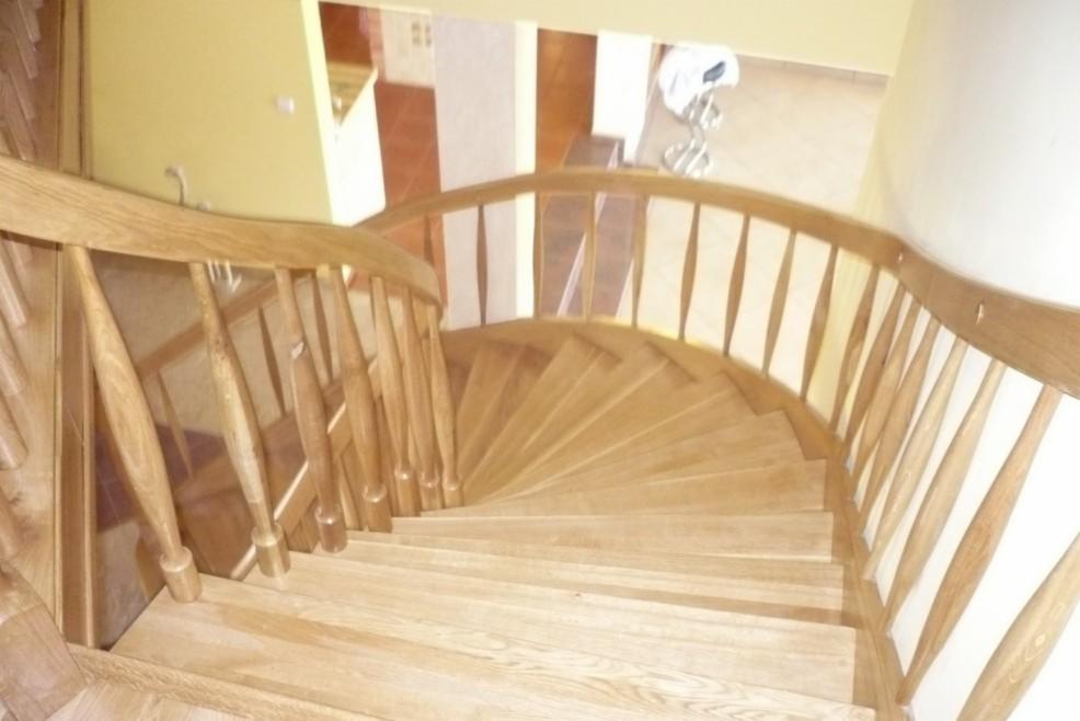 Spiralne schody drewniane z jasnego drewna, widok z góry, z delikatnym zakrzywieniem poręczy i prostymi pionowymi szczeblami, prowadzące w dół do innego pomieszczenia.