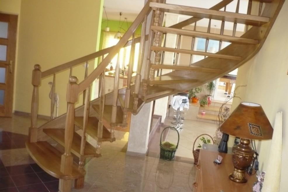 Drewniane schody kręcone z rzeźbionymi balustradami i ozdobną figurką ptaka, prowadzące na antresolę w jasnym wnętrzu domu.