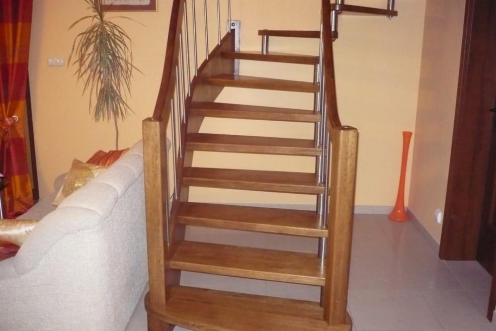 Drewniane schody kręcone z metalowymi barierkami w jasnym wnętrzu z beżowymi ścianami i szarą podłogą, obok widoczny fragment sofy.