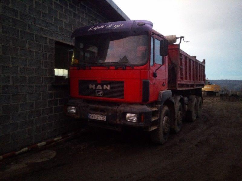 Czerwona ciężarówka MAN z otwartą skrzynią ładunkową, zaparkowana obok szarego, murowanego budynku. W tle widoczny krajobraz z koparką.