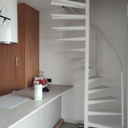 PAWI Patryk Wałach - Białe, kręcone schody metalowe w jasnym wnętrzu, widoczne z dołu, z otwartym otworem w suficie. Minimalistyczny design, szare płytki podłogowe i fragment blatu kuchennego.