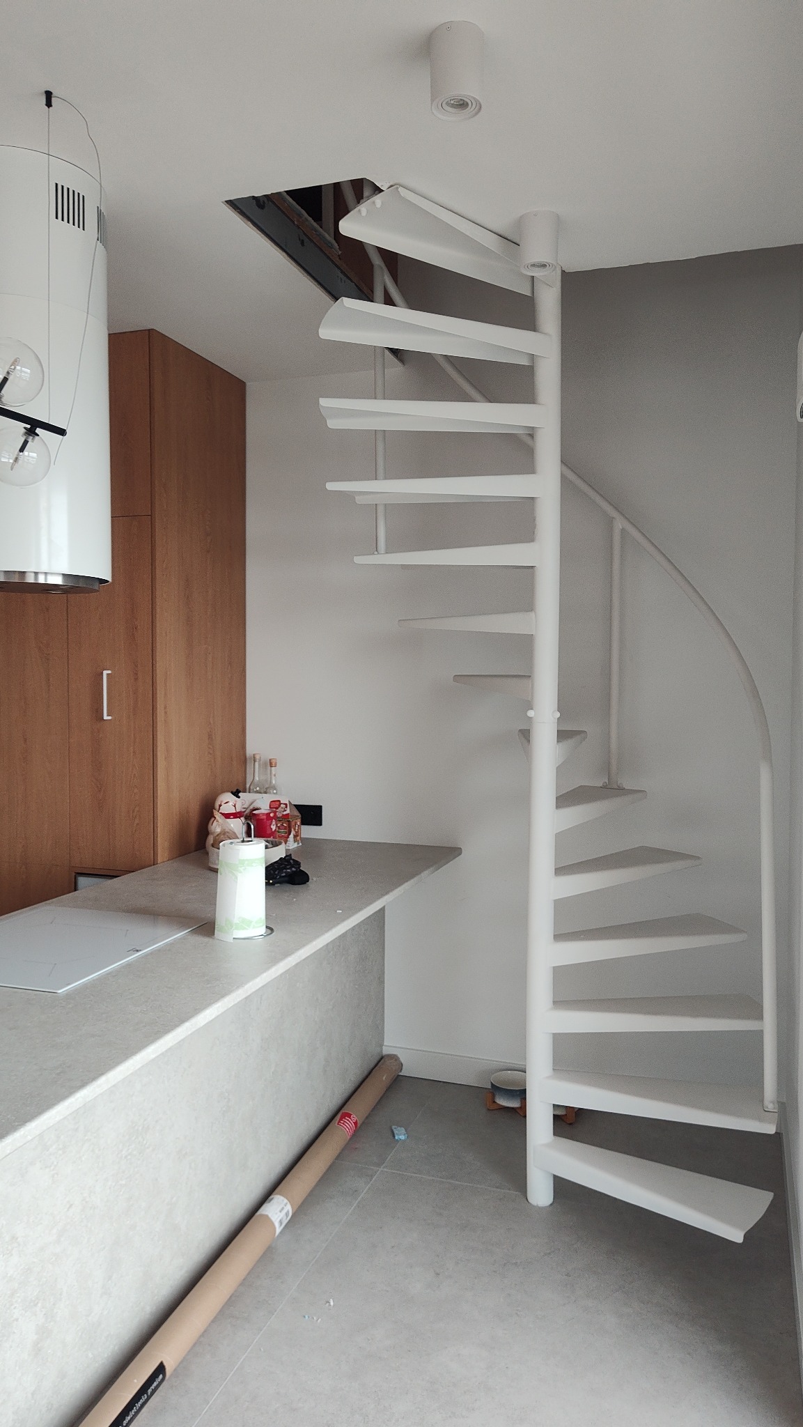 Białe, kręcone schody metalowe w jasnym wnętrzu, widoczne z dołu, z otwartym otworem w suficie. Minimalistyczny design, szare płytki podłogowe i fragment blatu kuchennego.