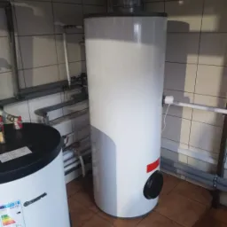 Bojler elektryczny stojący, biały, z czarnym panelem na dole, widoczny w kotłowni z rurami i kafelkami na ścianach.  Drugi bojler widoczny w tle po lewej.