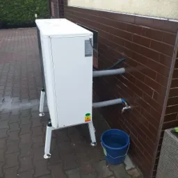 Biała pompa ciepła powietrze-woda, zamontowana na zewnątrz budynku z brązową ceglaną elewacją, widoczne szare rury podłączeniowe i niebieskie wiadro z wodą obok.