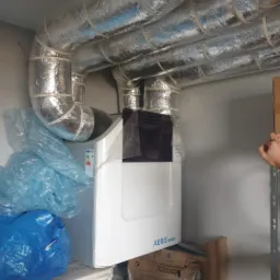 Biała jednostka rekuperatora Aeris Next z podłączonymi, zaizolowanymi przewodami wentylacyjnymi, widoczna w pomieszczeniu gospodarczym, częściowo zasłonięta plandeką i kartonami.