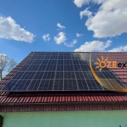 Panele słoneczne na dachu domu z czerwoną dachówką, błękitne niebo z chmurami w tle, widoczne logo firmy w prawym górnym rogu.