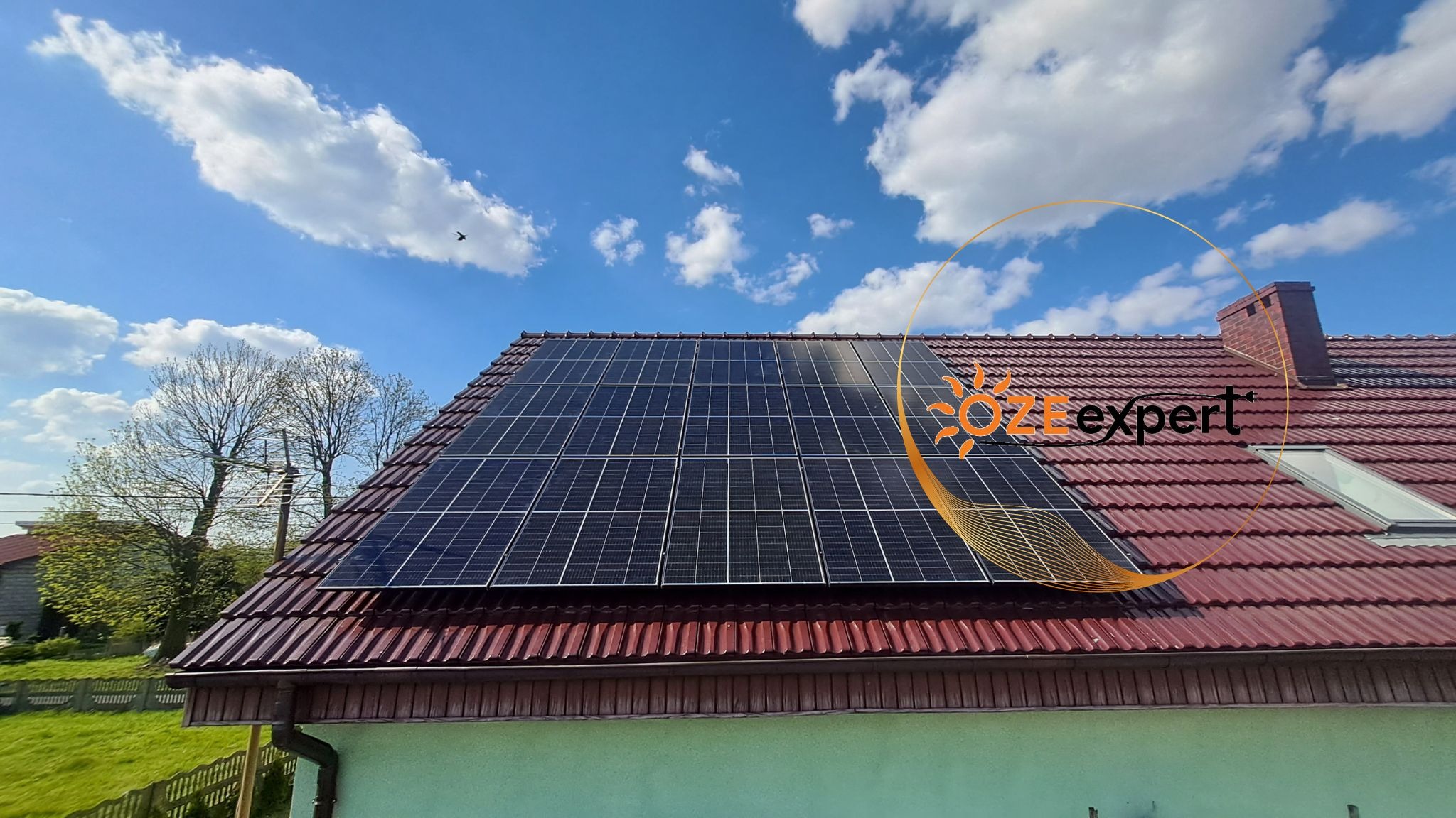 Panel słoneczny na dachu domu z czerwoną dachówką, błękitne niebo z chmurami w tle, logo firmy nałożone na zdjęcie.