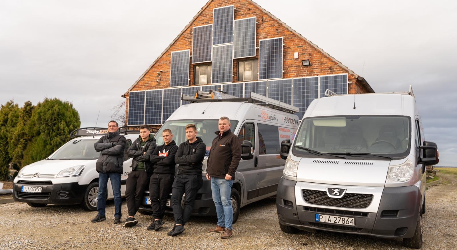 Grupa pięciu mężczyzn stoi na żwirowym podłożu przed ceglanym budynkiem z panelami słonecznymi na dachu, obok zaparkowane dwa busy z logo firmy zajmującej się instalacjami OZE.