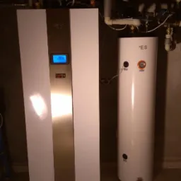 Biała, nowoczesna instalacja grzewcza z zbiornikiem buforowym i panelem sterowania, widoczne rury instalacyjne pod sufitem.