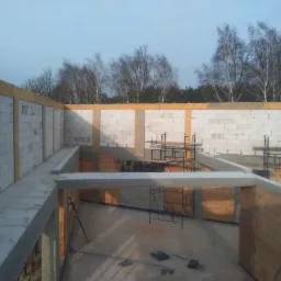 Wnętrze surowego stanu budynku z białych bloczków i fragmentami ścian z czerwonej cegły, widoczne rusztowania i elementy konstrukcyjne stropu z betonu, w tle drzewa bez liści pod błękitnym niebem.