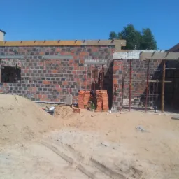 Wznoszenie ścian z czerwonej cegły w Kłecku, widoczne rusztowania i sterta cegieł, budynek w stanie surowym otwartym.