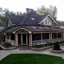 Dwukondygnacyjny dom z ciemną dachówką, ceglaną elewacją i werandą z białymi balustradami, otoczony zagospodarowanym ogrodem z trawnikiem i kamiennymi obrzeżami.