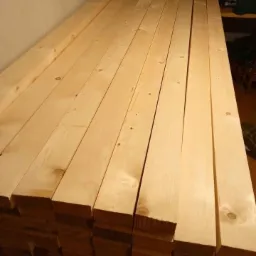 Stos jasnych, sosnowych desek ułożonych równo, widoczne sęki i naturalne usłojenie drewna, przygotowane do dalszej obróbki.