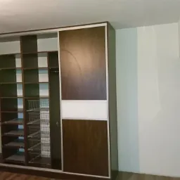 Wnętrze szafy przesuwnej z p&oacute;łkami i koszami drucianymi, widoczna część brązowych drzwi z białym pasem, obok telewizor na ścianie.