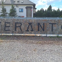 WERANTO - Mur z kostki brukowej z czarnym napisem 'WERANTO' w Sulejówku, zabezpieczony metalową barierką, na tle budynku i drzew.