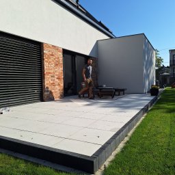 WERANTO - Nowoczesny taras z jasnych płyt betonowych obramowany ciemną kostką, mężczyzna przechadza się po tarasie, widoczny fragment domu z ceglaną wstawką i zielony trawnik.