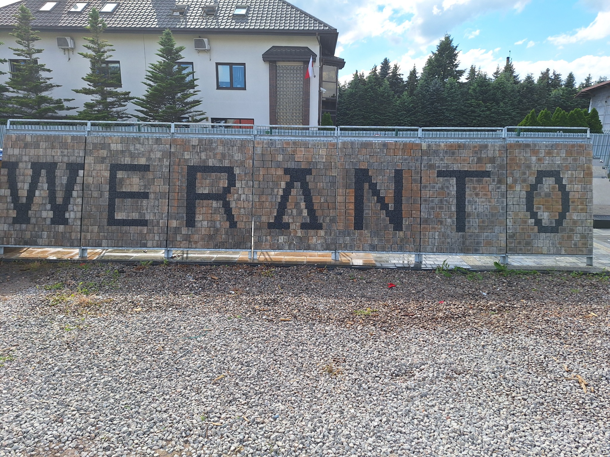 Mur z kostki brukowej z czarnym napisem 'WERANTO' w Sulejówku, zabezpieczony metalową barierką, na tle budynku i drzew.