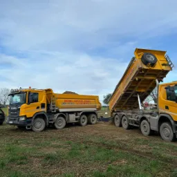 Dwa żółte samochody ciężarowe Scania na polu, jeden z podniesioną skrzynią ładunkową, z tyłu czarny samochód osobowy Volvo z otwartym bagażnikiem, błękitne niebo z chmurami w tle.