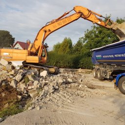 Firma Zdanpol - Pomarańczowa koparka gąsienicowa załadowuje łyżką pokruszony beton i gruz do niebieskiej ciężarówki; w tle zielona roślinność i dom.