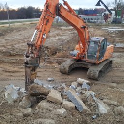 Firma Zdanpol - Pomarańczowa koparka z młotem hydraulicznym kruszy betonowe elementy na placu budowy, w tle składowisko kontenerów.