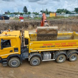 KZ Zdanpol Sp. z o.o. - Żółta wywrotka z napisem 'ŻWIROWNIA - PRACE ZIEMNE - ROZBIÓRKI' pod pełnym załadunkiem ziemi przez pomarańczową koparkę na budowie z widocznym wykopem pod fundamenty i pracownikiem w pomarańczowej...