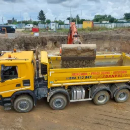 Żółta wywrotka z napisem 'ŻWIROWNIA - PRACE ZIEMNE - ROZBIÓRKI' pod pełnym załadunkiem ziemi przez pomarańczową koparkę na budowie z widocznym wykopem pod fundamenty i pracownikiem w pomarańczowej...