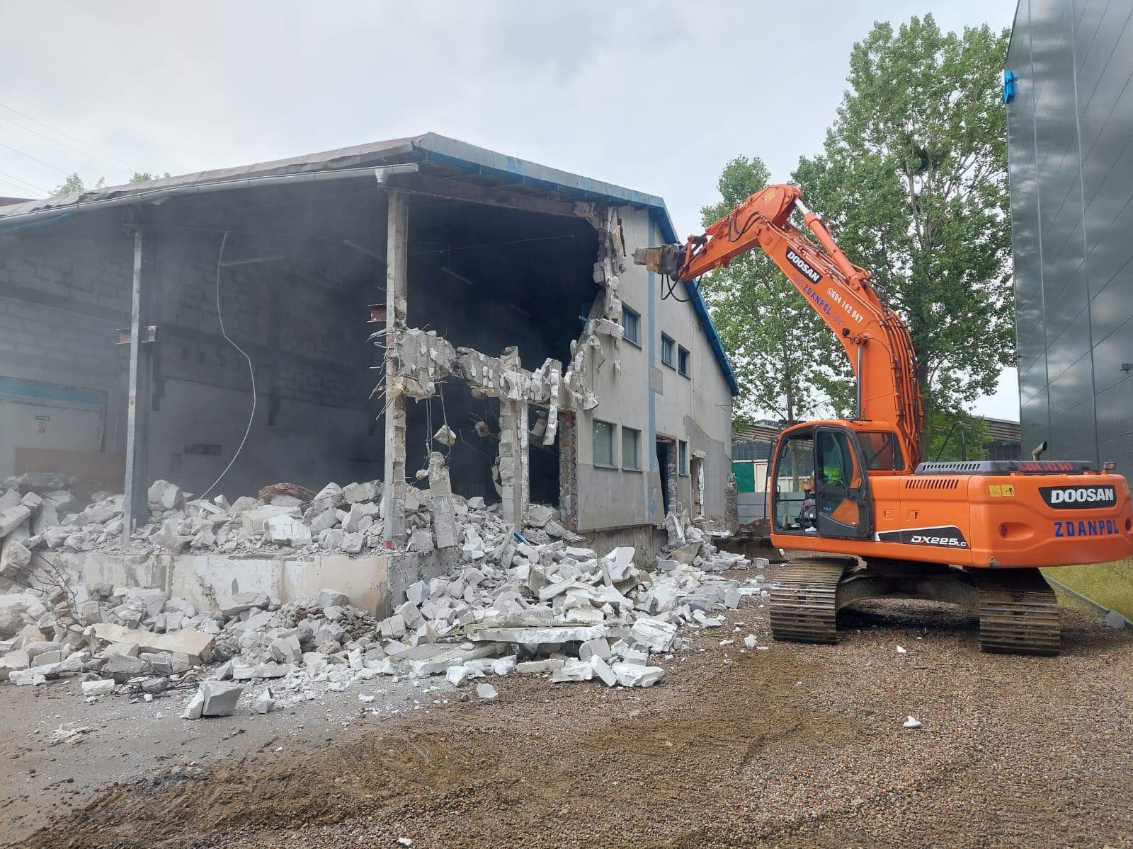 Pomarańczowa koparka Doosan z wysięgnikiem kruszy betonową ścianę budynku podczas rozbiórki, widoczne gruz i pył, budynek częściowo rozebrany.
