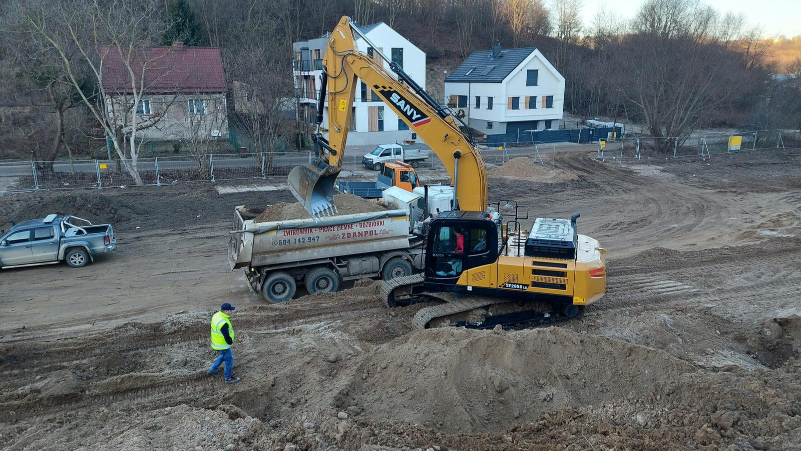Żółta koparka SANY nabiera łyżką ziemię i wsypuje ją do wywrotki na tle placu budowy z widocznymi budynkami mieszkalnymi i pracownikiem w żółtej kamizelce.
