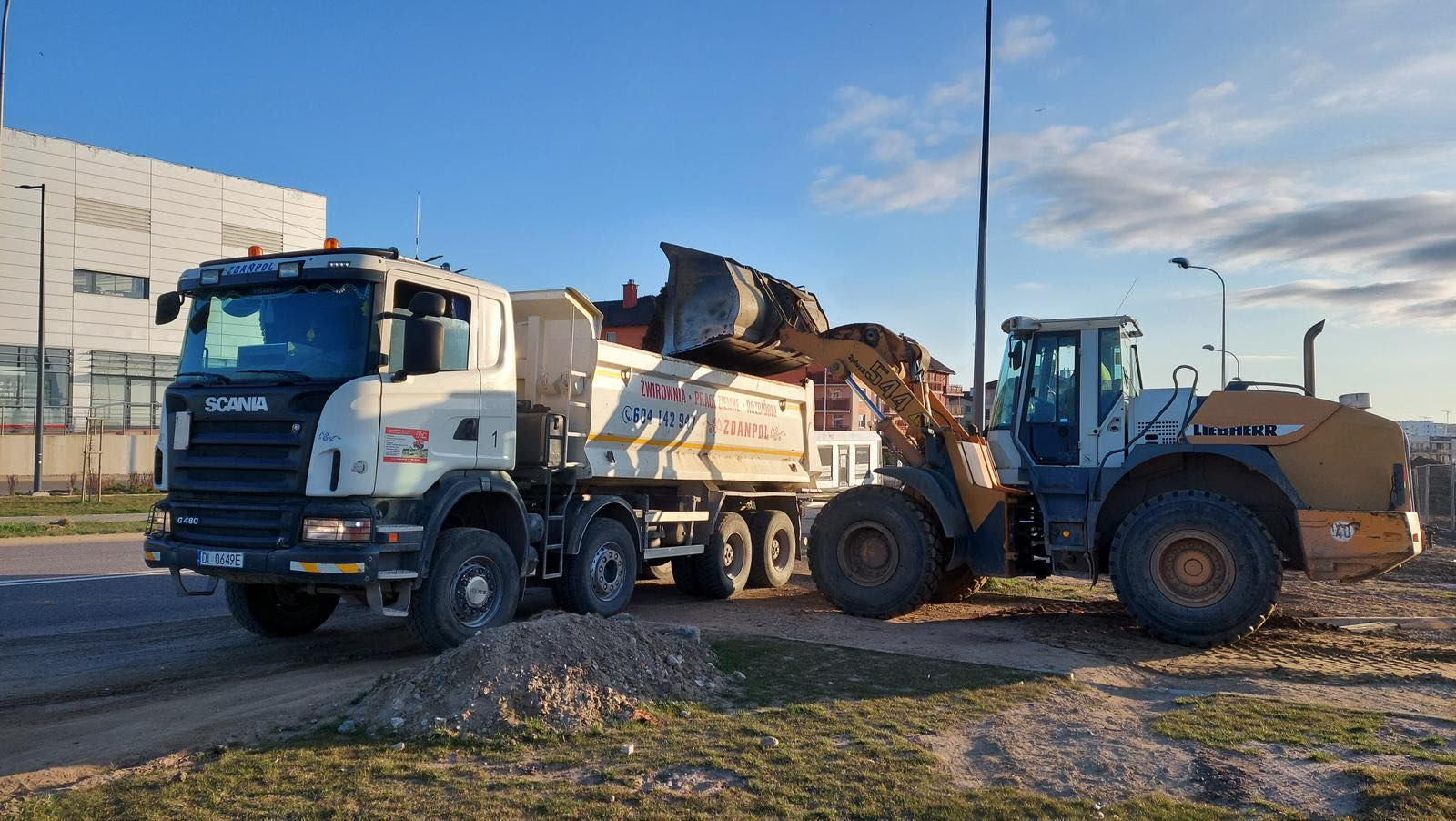 Żółta ładowarka Liebherr załadowuje materiał do białej ciężarówki Scania na placu budowy, widoczny niewielki pryzmat materiału.