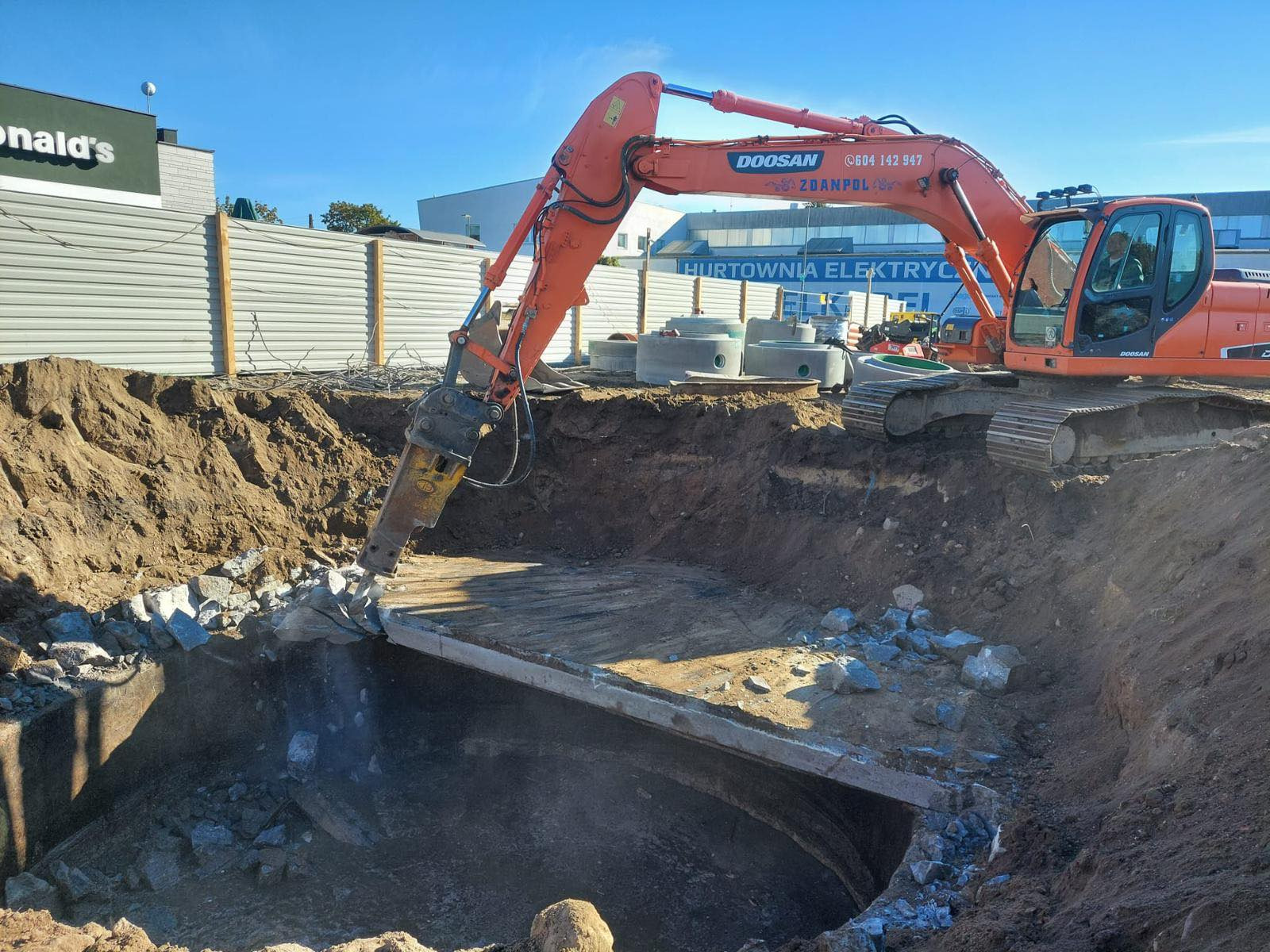 Pomarańczowa koparka z młotem hydraulicznym kruszy betonową płytę nad okrągłym wykopem, widoczne gruz i ziemia, w tle budynki i metalowe ogrodzenie.