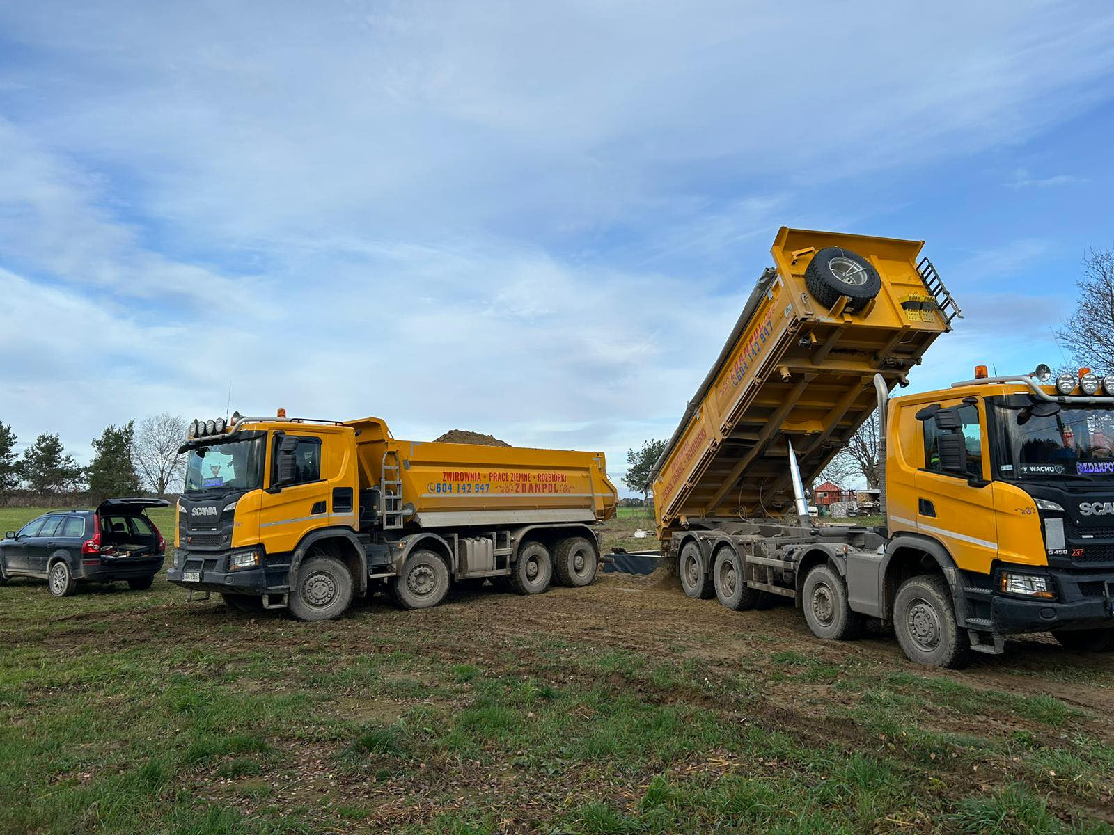 Dwa żółte samochody ciężarowe Scania na polu, jeden z podniesioną skrzynią ładunkową, z tyłu czarny samochód osobowy Volvo z otwartym bagażnikiem, błękitne niebo z chmurami w tle.