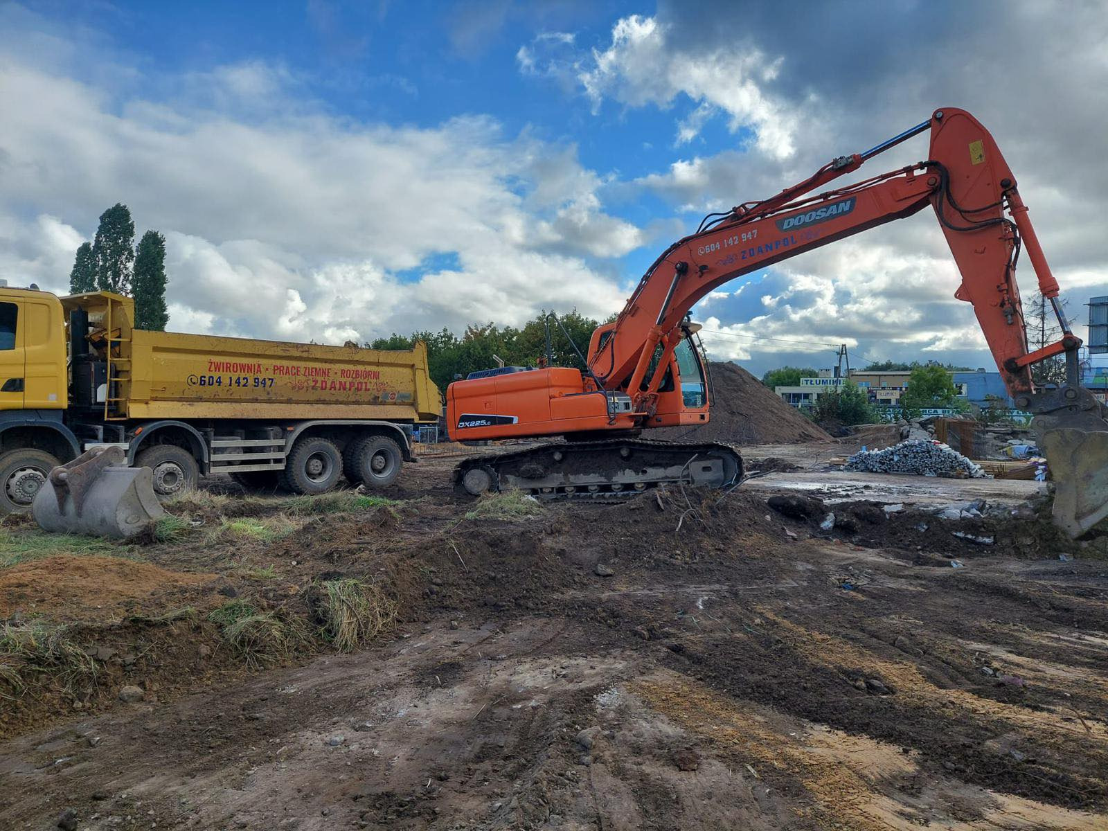 Pomarańczowa koparka Doosan DX225LC i żółta wywrotka z napisem 'Żwirownia - Prace Ziemne - Rozbiórki' na tle nieba z chmurami podczas prac ziemnych.
