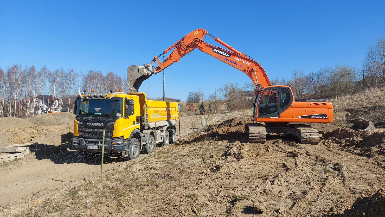 Pomarańczowa koparka Doosan nabiera łyżką ziemię i wysypuje ją do żółtej ciężarówki Scania na placu budowy w słoneczny dzień.