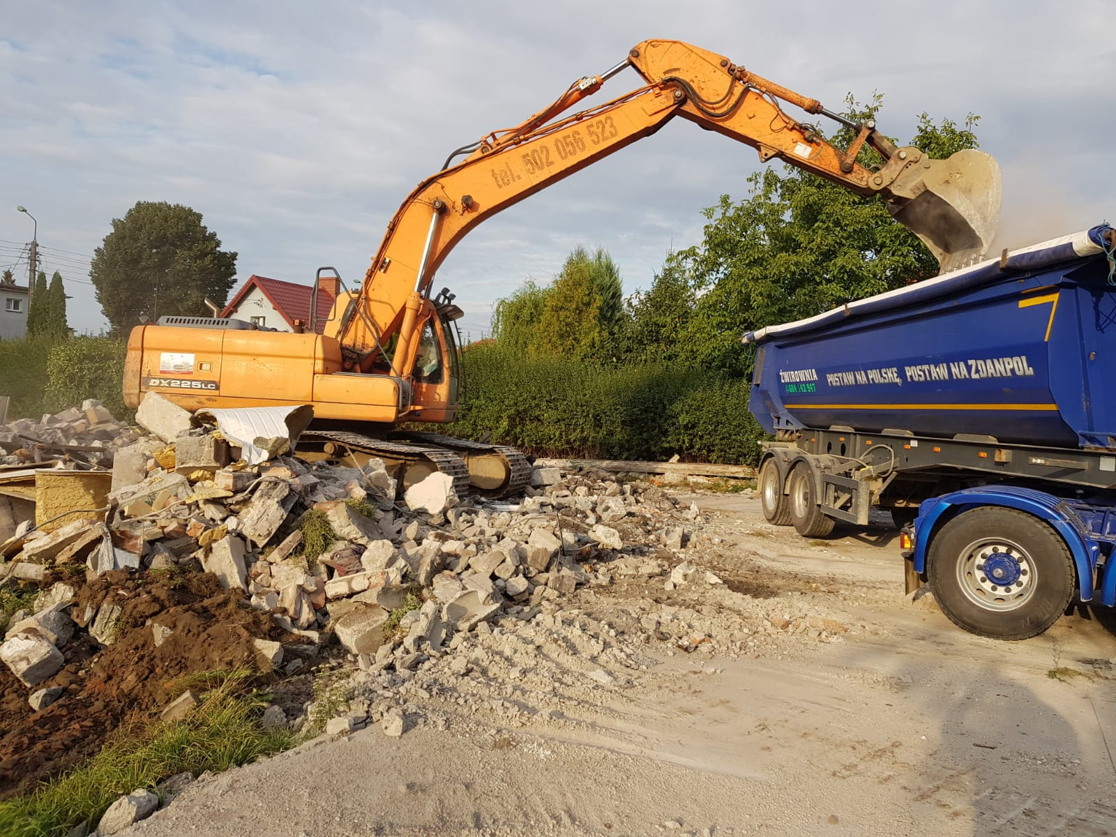 Pomarańczowa koparka gąsienicowa załadowuje łyżką pokruszony beton i gruz do niebieskiej ciężarówki; w tle zielona roślinność i dom.