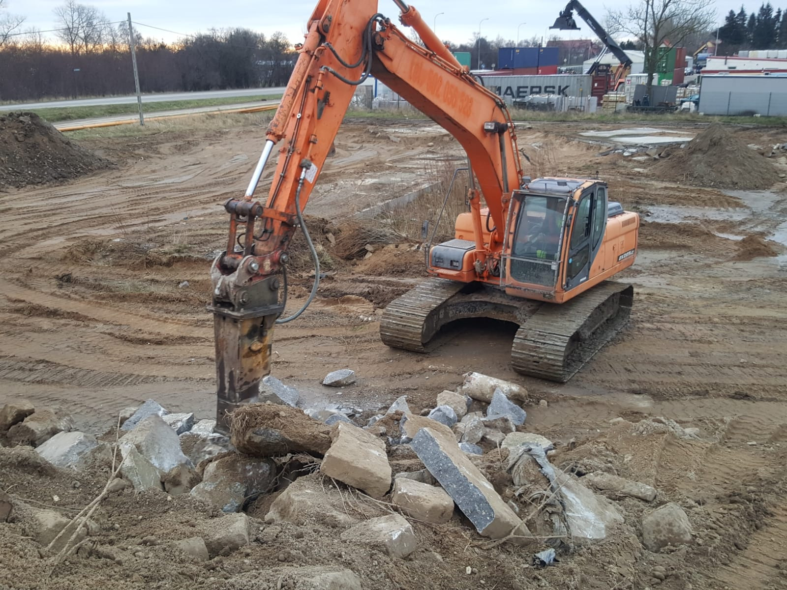 Pomarańczowa koparka z młotem hydraulicznym kruszy betonowe elementy na placu budowy, w tle składowisko kontenerów.