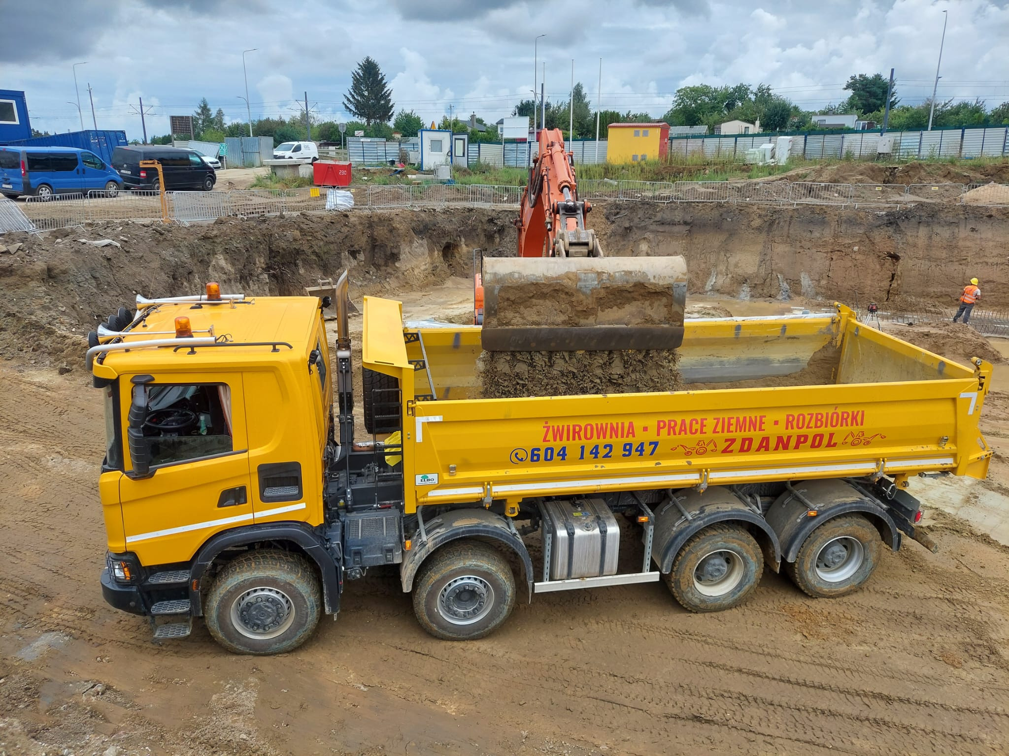 Żółta wywrotka z napisem 'ŻWIROWNIA - PRACE ZIEMNE - ROZBIÓRKI' pod pełnym załadunkiem ziemi przez pomarańczową koparkę na budowie z widocznym wykopem pod fundamenty i pracownikiem w pomarańczowej...