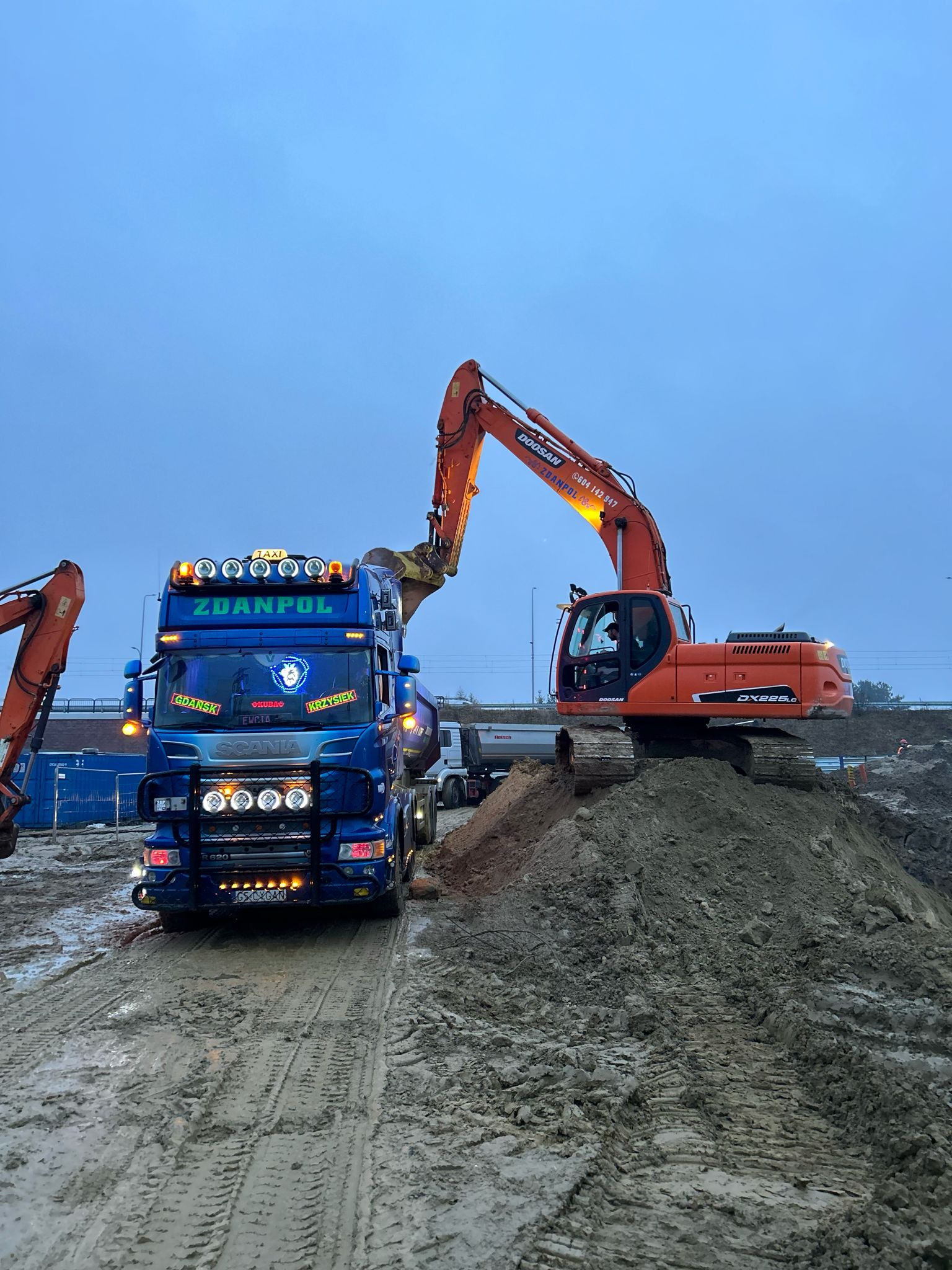 Pomarańczowa koparka Doosan DX225LC nabiera ziemię na wywrotkę Zdanpol Scania R620 w Kolbudach, w tle widoczny teren budowy.