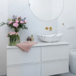 Nowoczesna łazienka z białą szafką pod umywalkę, marmurową umywalką nablatową, okrągłym lustrem i białymi płytkami w kształcie rybiej łuski, ozdobiona bukietami różowych kwiatów w wazonach.