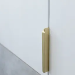 Detal zdjęcia białej szafki z minimalistycznym, pionowym uchwytem w kolorze szczotkowanego złota, widoczny fragment szarej podłogi.