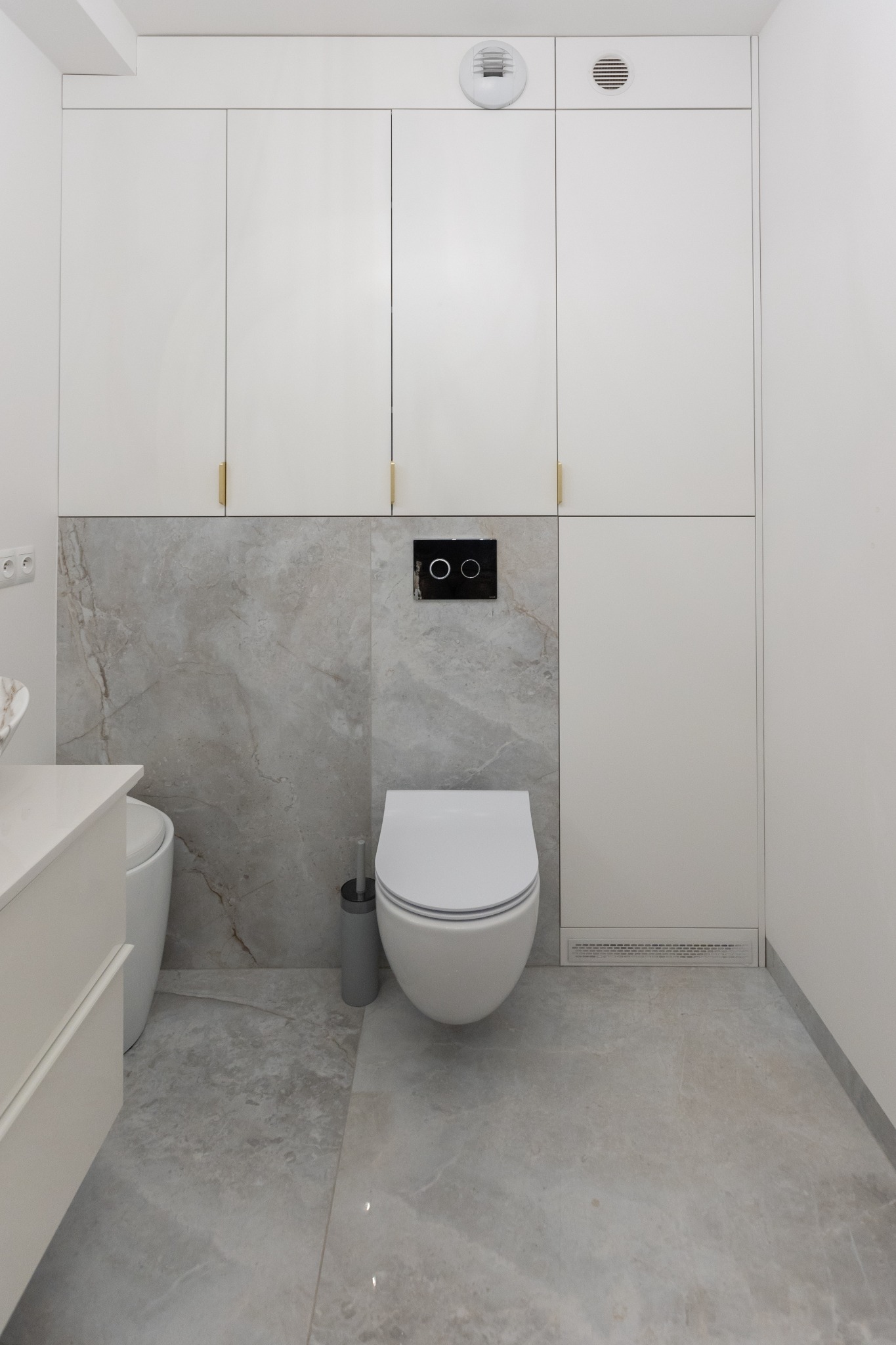 Nowoczesna łazienka w odcieniach bieli i szarości z podwieszaną toaletą, marmurową ścianą i szafkami w zabudowie.