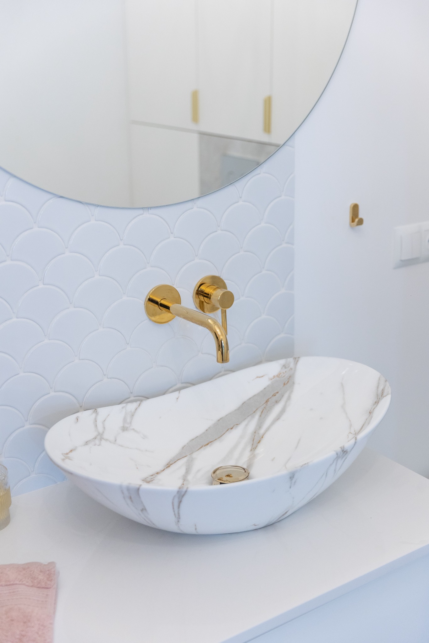 Nowoczesna łazienka z umywalką nablatową w stylu marmuru, złotą armaturą i białą mozaiką w kształcie rybiej łuski na ścianie.