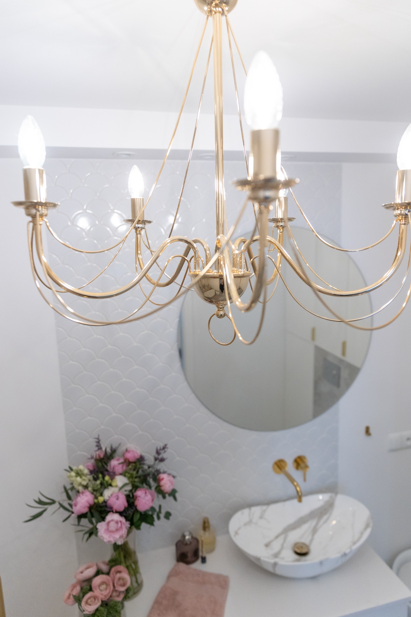 Elegancka łazienka z marmurową umywalką, złotą armaturą i dekoracyjnym, złotym żyrandolem na tle białych płytek o wzorze rybiej łuski oraz okrągłego lustra.