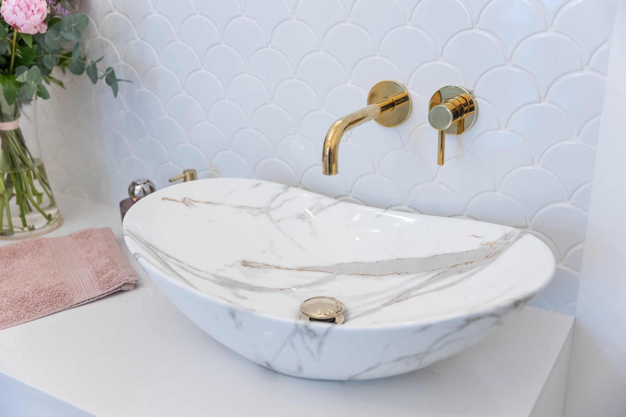 Biała marmurowa umywalka nablatowa o owalnym kształcie z złotą armaturą na tle ściany z białych płytek w kształcie rybiej łuski, z bukietem kwiatów w wazonie i różowym ręcznikiem obok.